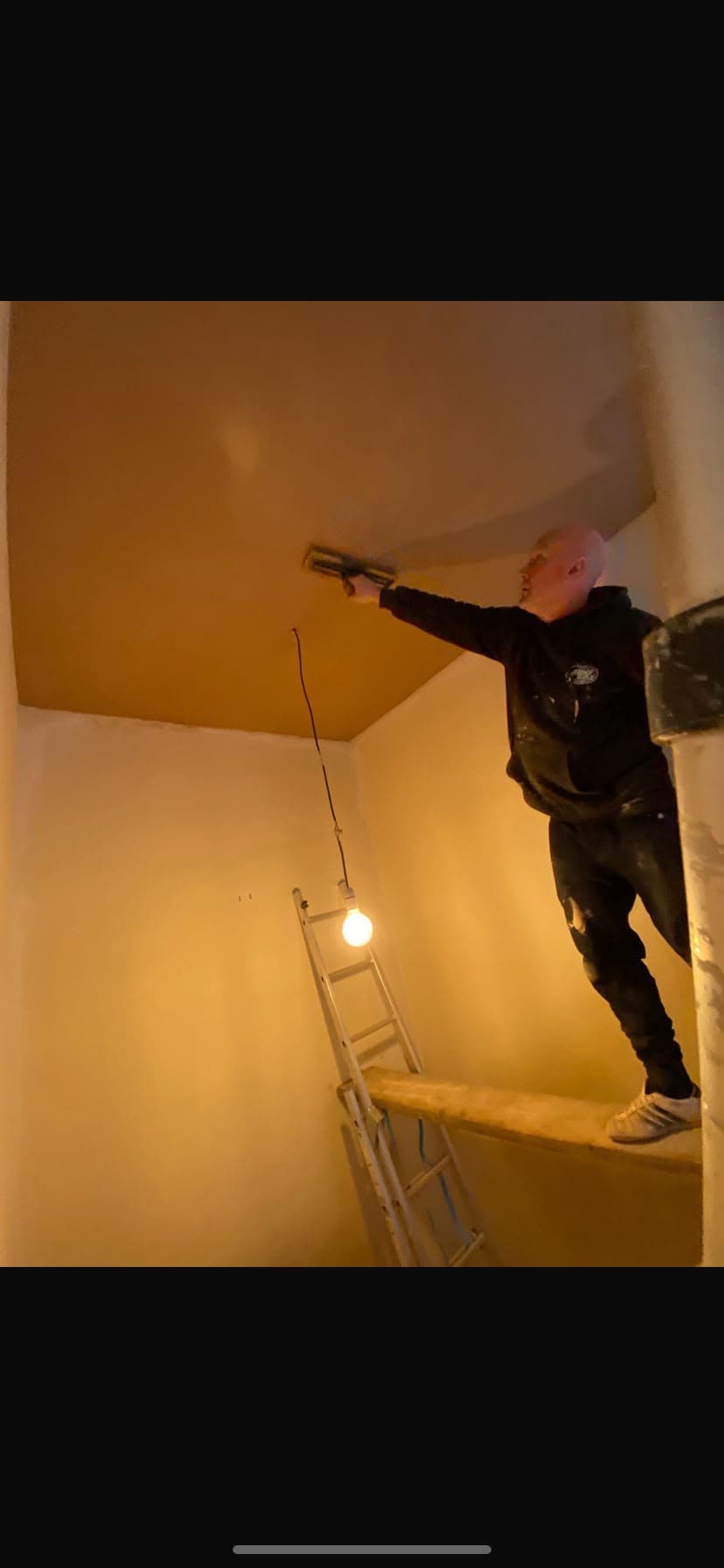 A plasterer working from a ladder and plank to skim a ceiling by lamplight